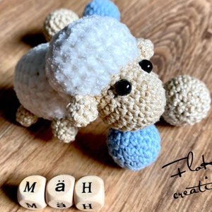 May include: A handcrafted crochet sheep with a fluffy white head and a beige body. The sheep has black eyes and is surrounded by small blue and beige crochet balls. The word "Mäh" is spelled out with wooden letter blocks.