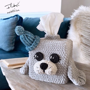 Puede incluir: Una funda para caja de pañuelos de tejido de punto gris en forma de foca que lleva un gorro azul de punto. La foca tiene ojos negros, una nariz negra y un hocico blanco.