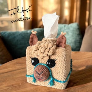 Puede incluir: Un cubridor de caja de pañuelos de tejido de punto en forma de llama con pelaje marrón, ojos azules y un detalle de cuerda azul y marrón. El cubridor de caja de pañuelos está sobre una mesa de madera.