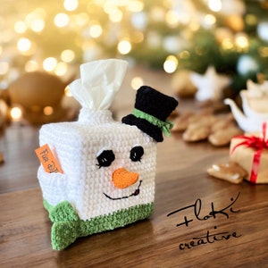 May include: A white crocheted snowman tissue box cover with a black top hat and green trim. The snowman has black eyes, an orange carrot nose, and a smiling mouth. A small orange tag reads "For D".