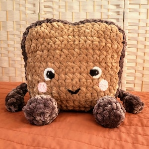Peut inclure: Un coussin en crochet marron en forme de carré avec un visage souriant et des joues roses. Le coussin a quatre petites pattes en crochet violet foncé.