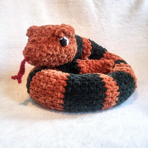 May include: A crocheted snake toy with a brown and black striped body and a red tongue. The snake has a black eye and is curled up in a circle.
