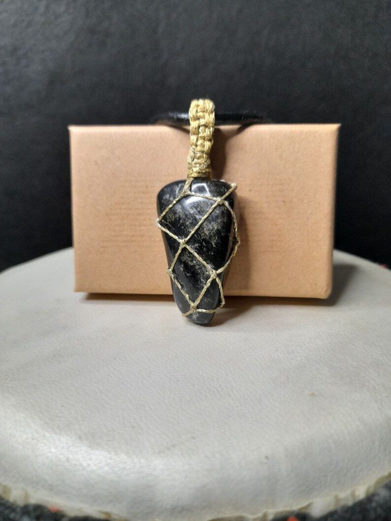 Amulet of Serpentine stone, uneven in shape, resembling a cone. The black color of the stone has a natural pattern of beige inclusions. The pendant is braided with golden waxed thread. The size is 4x2x1cm (high x top width x bottom width), weight 16g