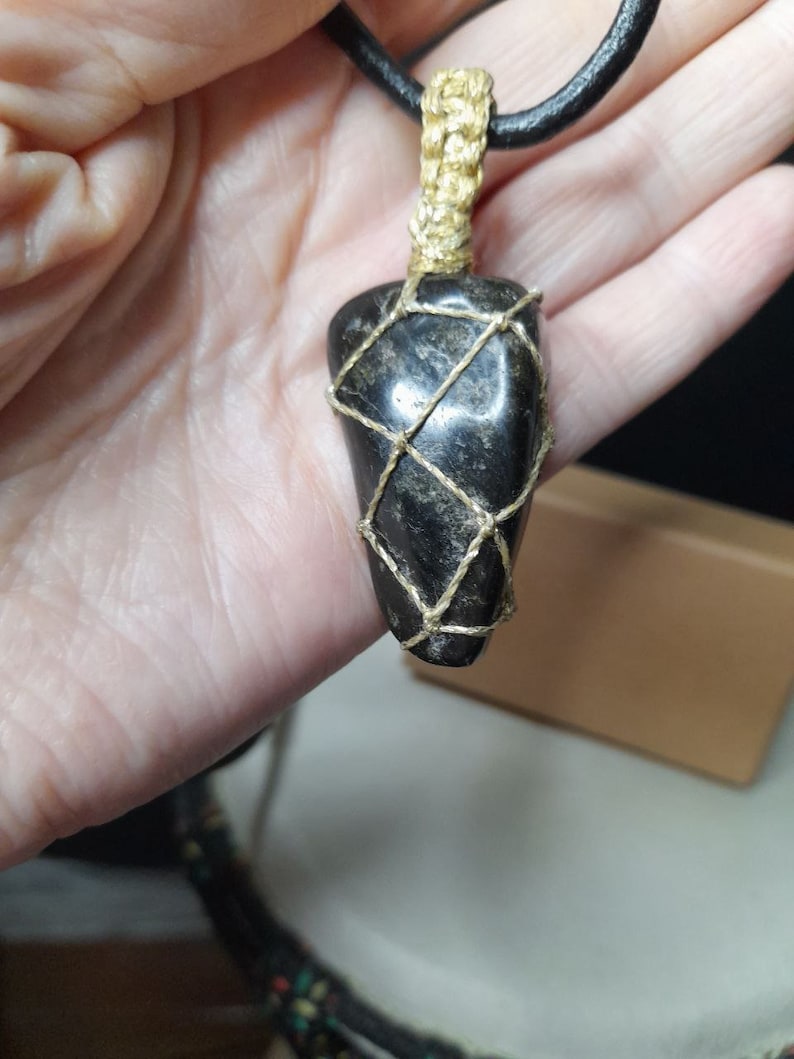 Amulet of Serpentine stone, uneven in shape, resembling a cone. The black color of the stone has a natural pattern of beige inclusions. The pendant is braided with golden waxed thread. The size is 4x2x1cm (high x top width x bottom width), weight 16g