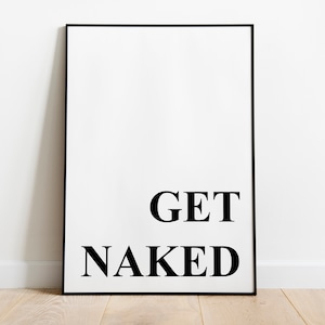 May include: A framed white print with the words "GET NAKED" in bold black letters. The artwork is displayed against a white wall and rests on a light-colored wooden floor. The frame is black.
