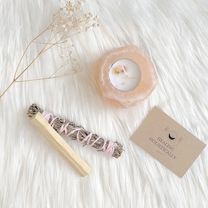 Puede incluir: Un portavelas de sal rosa del Himalaya con una vela blanca en su interior, un palo de salvia, un palo de palo santo y una pequeña tarjeta con el texto "Sanación holística".
