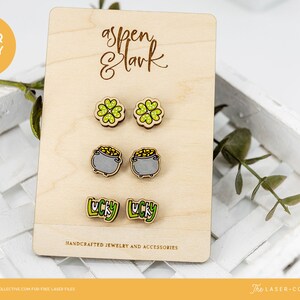 Op de afbeelding: Een set van drie paar lasergesneden houten oorbellen met een klavertjevier, een pot met goud en het woord "lucky". De oorbellen worden gepresenteerd op een houten kaart met de tekst "Aspen & Lark" en "Handgemaakte sieraden en accessoires".