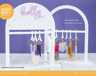 May include: White acrylic earring display stand with two different styles. The stand has a top shelf with holes for earrings and a lower shelf with hooks for hanging earrings. The stand is labeled "LASER READY" and "INCLUDES TWO DIFFERENT STYLES!"