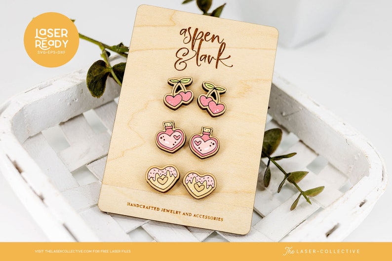 May include: A wooden sheet with the text "aspen & lark" and the text "HANDCRAFTED JEWELRY AND ACCESSORIES". The sheet displays six laser-cut wooden earrings: two cherry earrings, two heart-shaped potion bottle earrings, and two heart-shaped donut earrings.