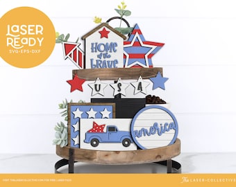 May include: A tiered wooden tray decorated with red, white, and blue patriotic themed wooden cutouts. The tray features a house cutout with the words "Home of the Brave", a truck cutout, a star cutout, and a round cutout with the word "america".
