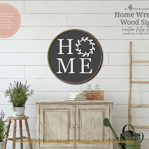 May include: A round wooden sign with a dark gray background and white lettering that reads "HOME" with a wreath around the "O". The sign is on a white shiplap wall with a wooden ladder and a watering can on a table.