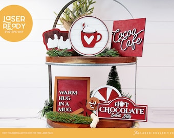 May include: A two-tiered wooden tray with a metal frame, decorated with red and white Christmas-themed wooden signs. The signs feature phrases like "Hot Chocolate Served Daily", "Warm Hug in a Mug", and "Cocoa Cafe".