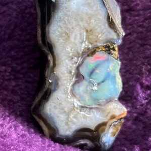 Puede incluir: Un colgante de oro con una piedra de ópalo natural engastada en un ágata negra. El ópalo muestra un arcoíris de colores, incluyendo rosa, azul y verde.