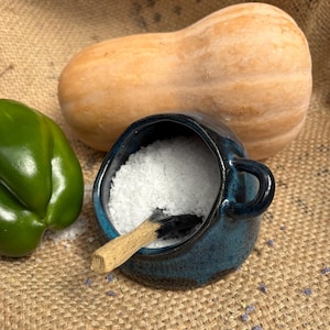 May include: A small, dark blue ceramic salt cellar with a handle, filled with white salt, and a wooden scoop. A butternut squash and a green bell pepper are in the background, all on a burlap surface.