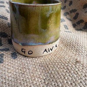 May include: A cylindrical ceramic mug featuring a glossy olive green glaze on the upper portion and a natural, unglazed beige base. The text "GO AWAY" is hand-stamped in black ink around the lower section of the mug, which has a smooth finish.