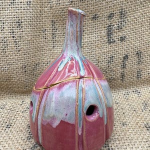 May include: A handmade ceramic garlic keeper, bulbous in shape with a slender neck. It's glazed in pink, blue, and white. The keeper has three holes on the side. The lid is pink with a gold accent. A decorative kitchen item.