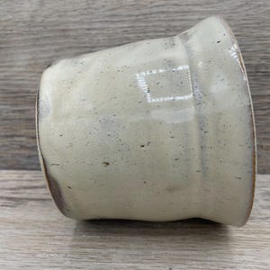 May include: A handmade ceramic mug with a light beige glaze and subtle speckled detailing. The mug has a slightly flared rim and a rounded base. The interior and rim have a darker brown colour. The mug is resting on a wooden surface.