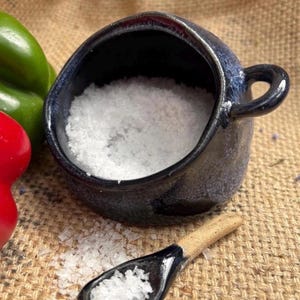 May include: A small, dark blue ceramic salt cellar filled with coarse white salt, with a matching spoon. The salt cellar has a small handle. Red and green peppers are visible in the background. The items are on a burlap surface.
