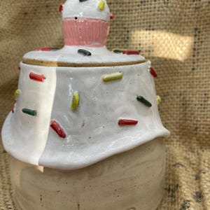 May include: A ceramic cupcake-shaped jar with a lid. The jar is off-white with white frosting and colourful sprinkles. The lid is topped with a pink cupcake and a red cherry. The jar is sitting on a burlap surface.