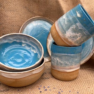 May include: A set of handmade ceramic tableware with a coastal theme. The set includes two bowls, two mugs, and two plates. The bowls and mugs have a light blue interior and a white and tan exterior. The plates have a light blue glaze.