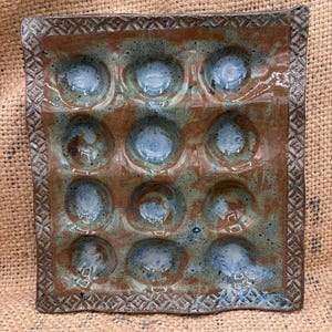 May include: Square ceramic egg tray with twelve circular indentations. The tray has a brown base with blue accents in the wells. The rim features a decorative geometric pattern. The tray is handmade and has a rustic appearance.