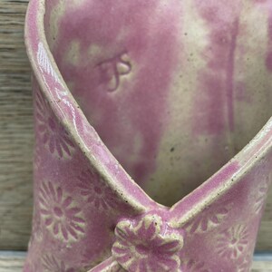 May include: A pink ceramic wall pocket with a floral pattern. The pocket is shaped like an envelope, featuring a flower detail and a small hole for hanging. The inside is a lighter shade of pink.