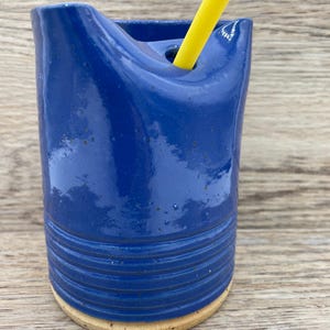 May include: A blue ceramic straw holder with a yellow straw sticking out of the top. The holder has a textured, ribbed design and a light brown base.