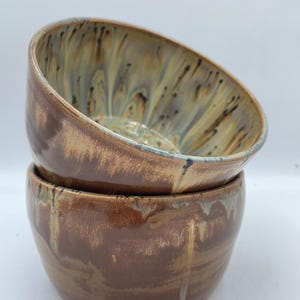 May include: Two ceramic bowls, one stacked on top of the other. The top bowl is a light brown with a white and blue marbled glaze. The bottom bowl is a darker brown with a white and gold marbled glaze.