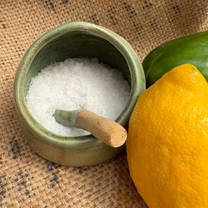 May include: A green ceramic salt cellar filled with coarse white salt, with a small wooden pestle. A bright yellow lemon and a green bell pepper are next to the salt cellar. The items are on a burlap surface.