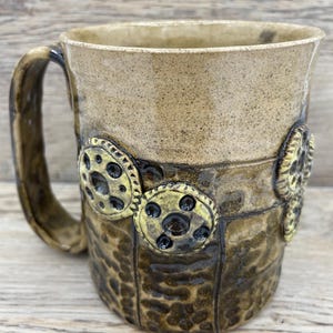 May include: A handmade ceramic mug with a tan upper section and a textured, dark brown lower section. The mug features decorative gear-shaped accents with black centres. The handle and rim are a darker brown. The mug is designed for hot drinks.