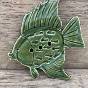 May include: A green ceramic fish ornament. The fish features detailed fins and scales, with three small holes in the body. This decorative item is likely for a coastal or nautical-themed home decor, adding a touch of marine life.
