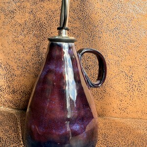 May include: A ceramic oil cruet with a glossy, deep purple and dark blue glaze. The vessel features a wide, rounded base that tapers towards the top, a curved handle on the side, and a silver-toned metal pour spout with a black plastic cap.