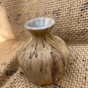 May include: A small, round ceramic vase with a narrow neck and flared rim. The vase has a mottled, earthy colour scheme with shades of tan, brown, and hints of blue around the rim. The vase is likely handmade.