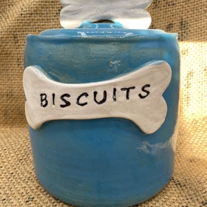 May include: A blue ceramic dog biscuit jar with a bone-shaped lid and a bone-shaped label that reads "BISCUITS". The jar is a rounded shape, ideal for storing dog treats. The jar is a vibrant blue colour, with white accents.