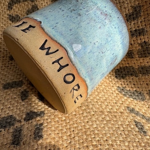May include: A ceramic mug with a speckled light blue glaze on the upper portion and a natural tan clay base. The text "THE WHORE" is stamped in black capital letters around the lower tan section of the mug, which has a cylindrical shape.