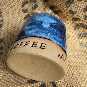 May include: A ceramic mug featuring a glossy, mottled blue glaze on the upper portion and a natural, unglazed beige base. The word COFFEE is stamped in black capital letters into the beige clay near the rim of the base, with partial text visible.