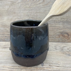 May include: A handcrafted ceramic utensil pot with a dark blue and brown glaze. A wooden spoon is placed inside, indicating its use for kitchen utensils. The pot's design is simple and rustic, suitable for a farmhouse or modern kitchen, measuring approximately 10 cm in height.