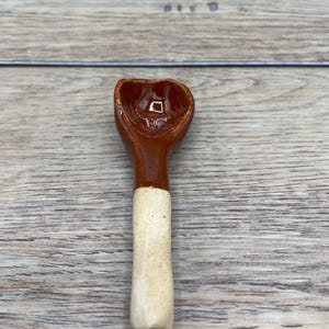 May include: A small, handmade ceramic spoon. It has a brown bowl and a cream-coloured handle. The spoon is designed with a rounded bowl and a long, slender handle. The ceramic spoon is likely used for serving spices or condiments.