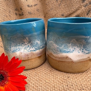 May include: Two ceramic mugs with a coastal design. The top of the mugs are a bright blue, transitioning to white and tan, resembling a beach scene. A vibrant red flower is in the foreground.