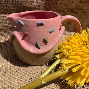 May include: A ceramic creamer jug with a pink glazed top section featuring small, raised, multi-coloured sprinkles and a smooth, speckled tan base. The jug has a curved handle and a pouring spout, positioned next to a bright yellow flower.