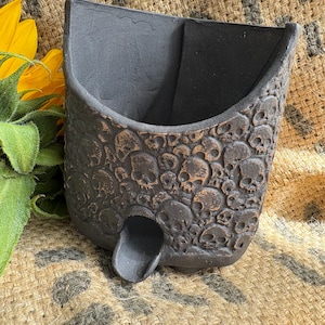May include: A matte black ceramic wall planter featuring a textured surface covered in a repeating embossed skull pattern. The vessel has a wide, open top with a curved rim and a small, protruding drainage spout located at the bottom centre.