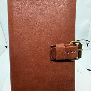 May include: A brown leather journal with a buckle closure. The journal has a textured surface and a brass-colored buckle. The leather appears to be aged, with some visible wear and tear. The journal is standing upright on a black metal stand.