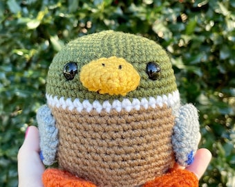 Puede incluir: Un juguete de peluche de pato tejido a crochet con hilo verde, marrón, naranja, blanco y gris. El pato tiene ojos negros y un pico amarillo.