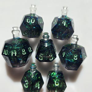 May include: Set of seven polyhedral dice, each shaped like a potion bottle. The dice are dark with green and blue glitter, and white numbers. The dice are for tabletop games.