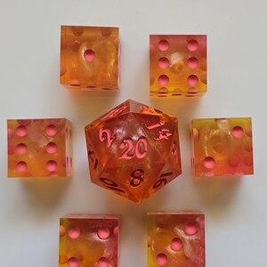 Puede incluir: Un juego de siete dados de resina en tonos naranja, amarillo y rosa. El juego incluye seis dados de seis caras y un dado de veinte caras, todos con puntos rosas. Los dados tienen un aspecto veteado.