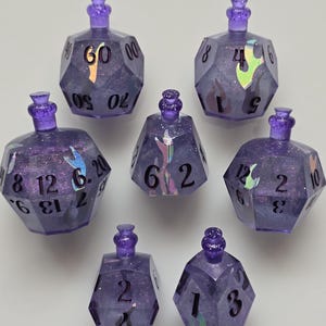 Puede incluir: Siete dados morados de múltiples caras con purpurina y acentos iridiscentes. Cada dado tiene un pequeño tapón decorativo y está numerado con números blancos. Los dados están diseñados para juegos de mesa.