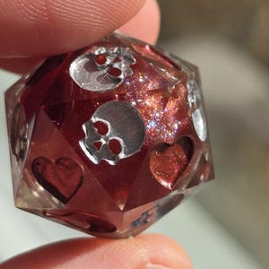 May include: A faceted, multi-sided die with a red interior and silver skull and heart designs. The die is held between two fingers, showcasing its intricate details and sparkling inclusions. A unique gaming accessory.
