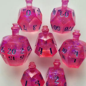 Puede incluir: Un conjunto de siete dados poliédricos rosas con purpurina, cada uno con forma de botella de poción. Los dados tienen números azules y están diseñados para juegos de rol de mesa. Los dados están dispuestos sobre una superficie blanca.