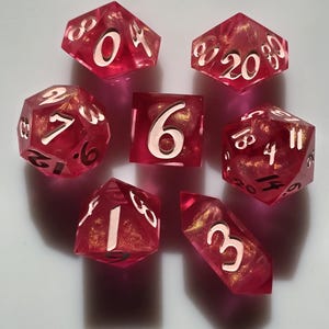 May include: Set of seven pink and gold dice with white numbers. The dice are in various shapes, including a d4, d6, d8, d10, d12, and d20.