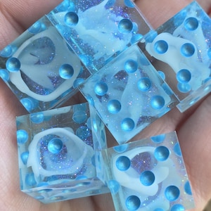 May include: Six clear resin dice with a blue and white design. Each die has blue dots and a white, swirling design inside. The dice are held in a hand, and the overall effect is a cool, aquatic theme.